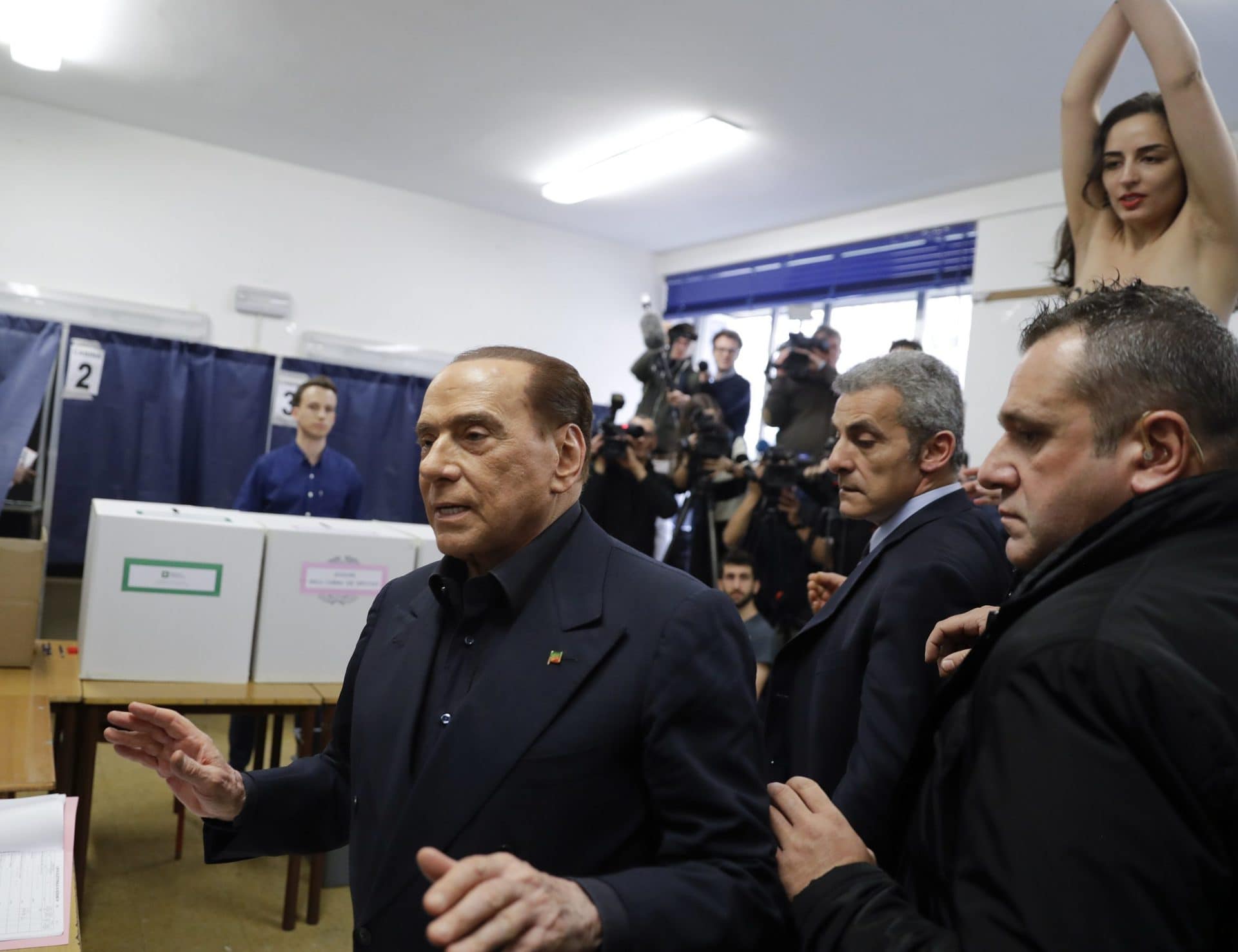 Silvio Berlusconi, left, arrives to vote as a bare-breasted woman protests in background. AP Photo/Luca Bruno