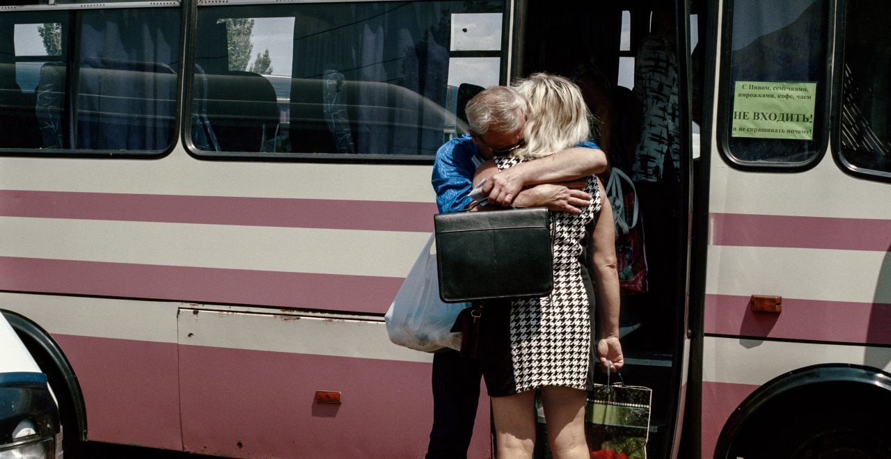 Image of a man and woman hugging in front of a bus. Image description: A bus departing from Kurakhovo, a frontline town, to the entry checkpoint into the non-government controlled Donetsk. After reaching the checkpoint, passengers will have to queue for many hours, in the middle of a mine field, waiting to cross. Despite harsh conditions, over a million people cross this line every month. One of the major reasons for traveling is to visit family members on the other side. Photo by Anastasia Taylor-Lind. Caption by Alisa Sopova..